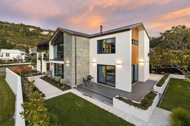 Contemporary multi-level residential house on a landscaped suburban hillside site, featuring white rendered walls, a prominent grey stacked-stone facade, timber-clad vertical sections, dark-framed glazing, and a low-pitched gabled roof. Paved terraces, raised white garden beds, manicured lawns, and clipped planting surround the home.