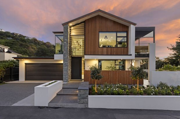 A contemporary two-storey house viewed from the street at dusk, featuring a pitched gable roof, vertical timber-look cladding, dark stone detailing, large glazed openings, a sheltered upper balcony, attached garage, and landscaped front garden with rendered planter walls.