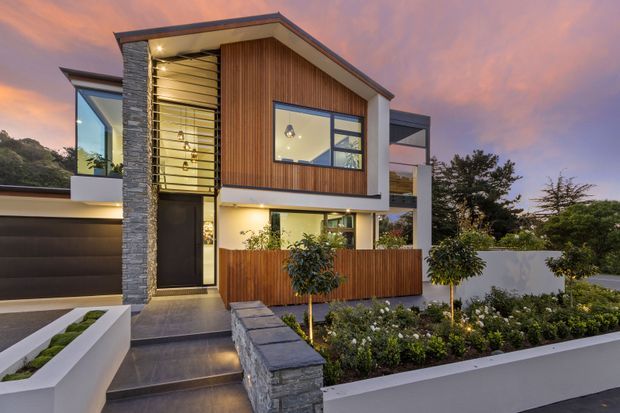 Contemporary multi-storey residence photographed at dusk, featuring a flat roof, strong geometric volumes, vertical timber cladding, stacked-stone walls, expansive glazing, and a landscaped front garden. A tall glazed entry volume with horizontal sun-shading elements forms a prominent architectural feature.