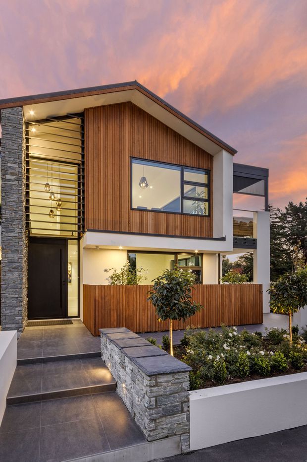 Two-storey contemporary residential facade at dusk, featuring a steep asymmetrical gabled roof, vertical timber cladding, dark stone veneer, large black-framed glazing, and a recessed illuminated entry. A landscaped front garden is enclosed by timber and stone walls, with a covered upper-level outdoor area visible to the right.