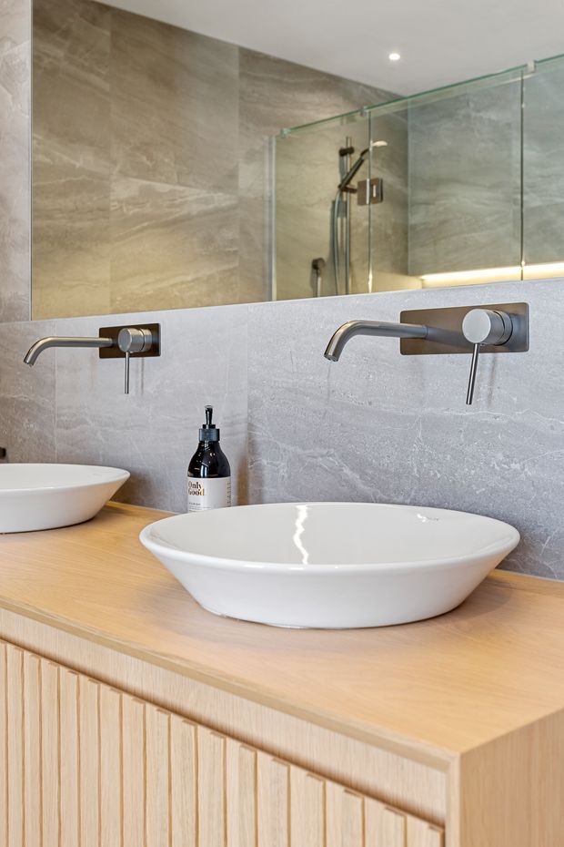 Contemporary bathroom vanity with two white vessel basins on a light timber fluted cabinet, wall-mounted brushed metal taps, pale stone-look wall surfaces, and a large mirrored backsplash. A glass-enclosed shower with matching stone-look tiling is visible in the reflection.
