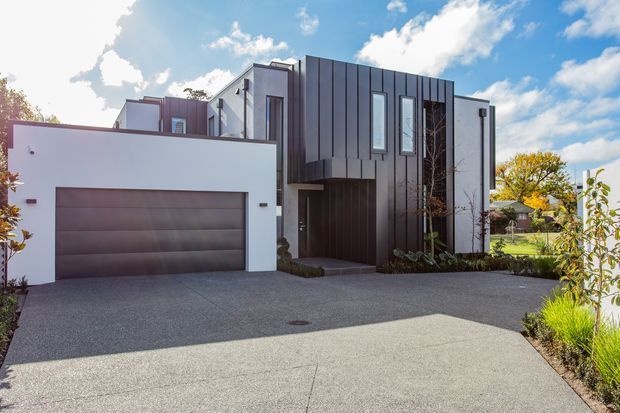 Contemporary two-storey residential home viewed from the front, featuring a clean white rendered garage wing, a dark vertically clad central facade, narrow vertical windows, recessed entry, and a broad concrete driveway with landscaped planting.