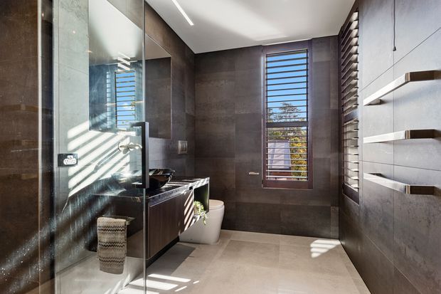 Contemporary bathroom with a walk-in glass shower, dark stone-look wall surfaces, a floating dark timber vanity with black basin, wall-mounted toilet, and open timber shelves. Horizontal louvre windows bring in strong natural light, while large pale floor tiles balance the darker finishes.