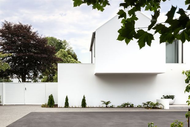A minimalist white residential façade viewed from the driveway, featuring clean geometric volumes, a projecting upper storey, narrow vertical windows, a white boundary wall and gate, and restrained planting. Mature trees frame the property.