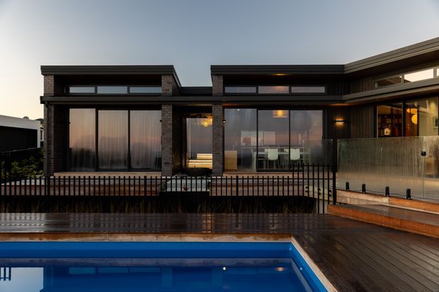 Exterior view of a contemporary multi-level residence at dusk, featuring dark brick and dark cladding, extensive floor-to-ceiling glazing, a timber deck, metal balustrades, and a swimming pool in the foreground. Warm interior lighting is visible through the windows.