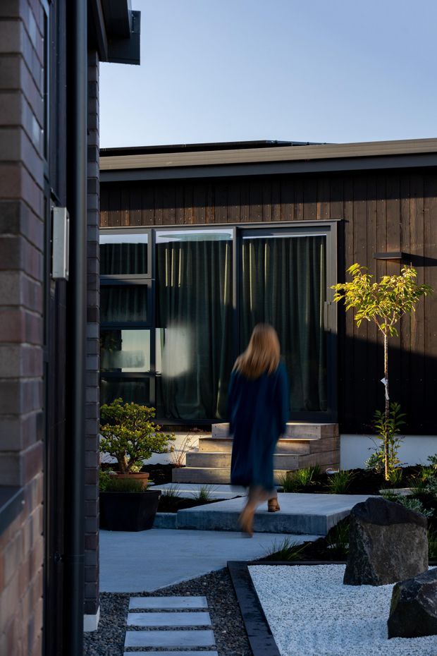 A contemporary courtyard viewed between dark brick and timber-clad building volumes. The landscaped outdoor space features concrete paths with stepping stones, white gravel, large boulders, clipped planting, a small tree, and timber steps leading to large glazed doors with dark curtains. A blurred person walks through the courtyard.