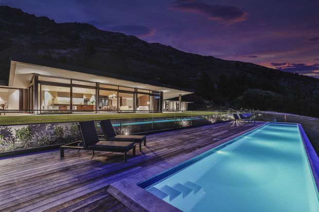Twilight exterior view of a sleek, low-profile contemporary home set against a rugged mountain landscape. Extensive floor-to-ceiling glazing opens onto a broad timber pool deck, with a long illuminated swimming pool, integrated entry steps, and minimalist outdoor lounge furniture in the foreground.