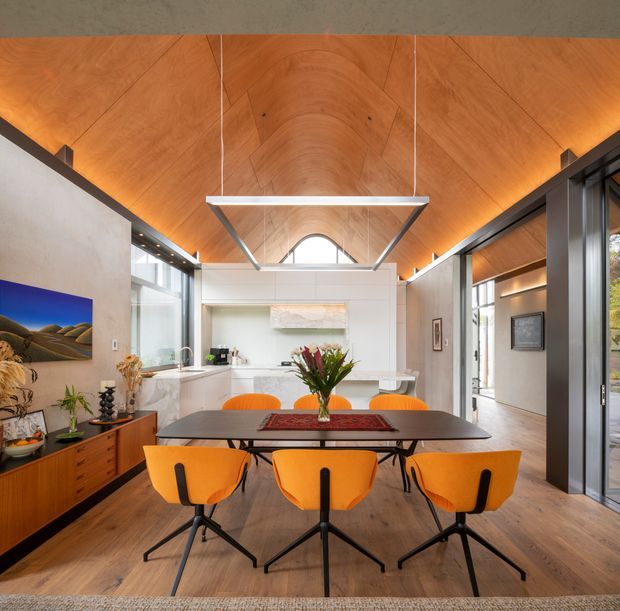 A contemporary dining area in a striking vaulted interior, centred on a dark rectangular dining table with six vivid orange chairs. Warm timber panels line the barrel-vaulted ceiling and timber flooring, while large glazed openings, white cabinetry, concrete-toned walls, and a geometric suspended light create a bright, refined atmosphere. A small arched window and connected living areas are visible beyond the dining space.