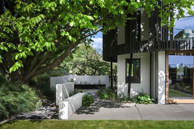 Exterior view of a contemporary two-storey residence framed by mature landscaping. A dark upper volume with vertical cladding projects over a light brick lower level, with extensive glazing, a sheltered entry, paved paths, built-in white masonry seating or retaining walls, and lush planting beneath a broad tree canopy.