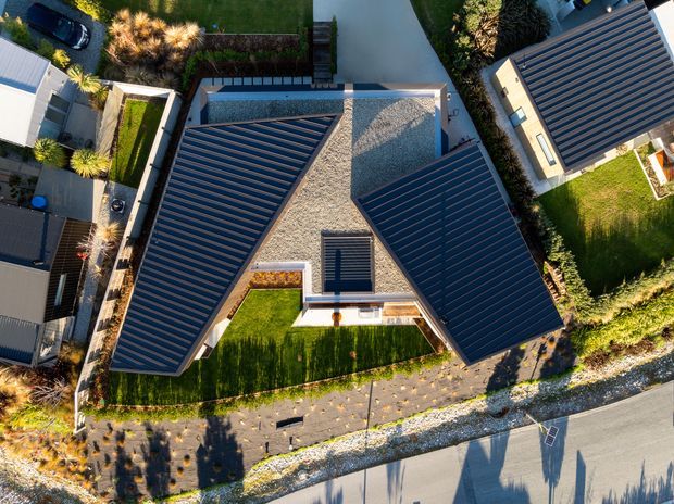Aerial top-down view of a modern residential property with a distinctive branching roof plan. Dark metal pitched roof sections surround a central light-coloured gravel or textured roof area, with landscaped lawns, planted garden beds, paving, and neighbouring homes visible around the site.