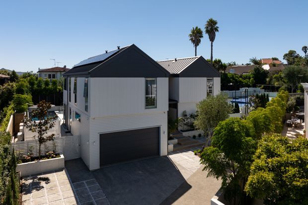 Exterior view of a contemporary multi-level suburban residence with dark gabled metal roofs, crisp white horizontal cladding, large windows, and an integrated double garage. Paved paths and driveway, landscaped gardens, neighbouring homes, and a partially visible backyard pool are present.