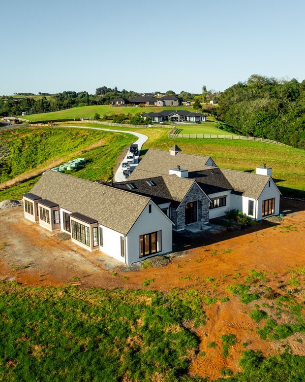 Aerial view of a newly built single-storey rural residence with multiple gabled wings, white rendered walls, dark stone accents, and grey textured pitched roofs. The home sits on a large landscaped and partially undeveloped property, with a curved driveway, fencing, expansive green fields, mature trees, and neighbouring homes in the distance.
