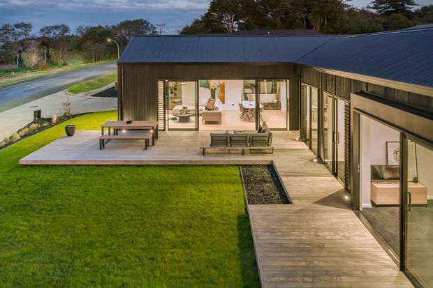 A low-profile contemporary house viewed from the landscaped exterior, with dark vertically clad walls and pitched metal roofs arranged around a timber-decked courtyard. Large glazed sliding doors open to a bright living area, while lawn, paving, and a gravel drainage strip frame the outdoor space.