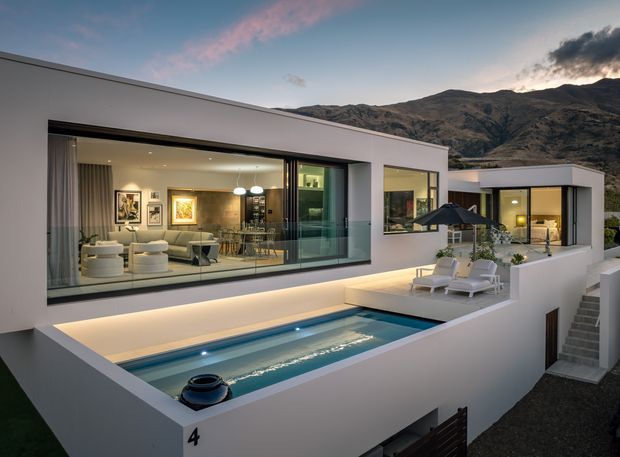 Contemporary flat-roofed white villa built into a sloping site, photographed at dusk with rugged mountains behind it. The view centres on a narrow rectangular swimming pool, paved terrace with lounge chairs and umbrellas, and expansive floor-to-ceiling glazing revealing a warm, art-filled open-plan living and dining area. Clean linear geometry, integrated lighting, and pale rendered walls create a refined indoor-outdoor composition.