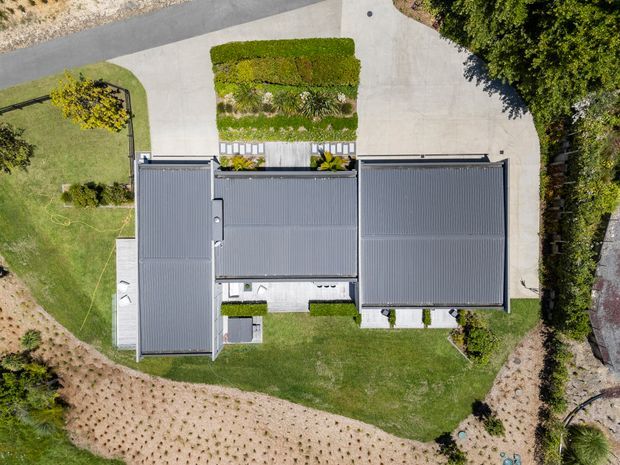 A top-down aerial view of a contemporary residential property, showing several connected dark grey metal-clad roof volumes arranged around landscaped garden areas. The site includes lawns, clipped hedges, planted garden beds, concrete paths and driveway areas, with surrounding vegetation and a partially visible road.