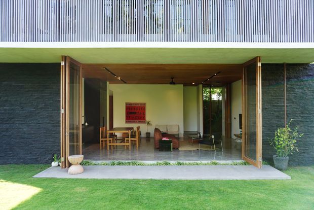 Exterior view of a contemporary pavilion-like residence opening onto a manicured lawn. Large timber-framed bi-fold or folding glass doors are fully open, revealing a compact combined living and dining area with timber ceiling, polished concrete-like floor, dark stone-textured walls, simple furniture, and track lighting. A green flat roof or fascia projects over the opening, while vertical metal screening is visible above.