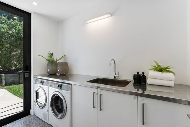 Bright, compact modern laundry room with a white fixed cabinetry run, stainless-steel benchtop and integrated sink, front-loading washer and dryer, and minimalist black hardware. A full-height glazed door opens to a garden courtyard, while potted plants, folded towels and black containers add restrained decorative touches. The room has a clean, practical and contemporary feel.
