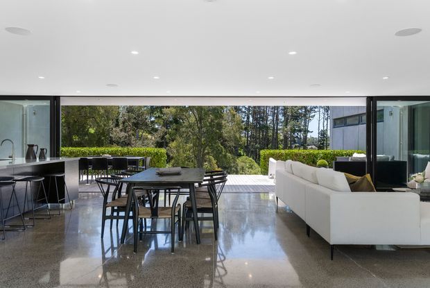 Open-plan contemporary living, dining, and kitchen space with full-width retractable or glazed openings connecting directly to a landscaped outdoor terrace. The room features a polished grey concrete floor, white ceiling with recessed downlights, dark-framed glazing, a white sectional sofa, and a dark dining table with black chairs. A linear kitchen with dark cabinetry and a small island is visible on the left. The space is bright and strongly connected to the garden.