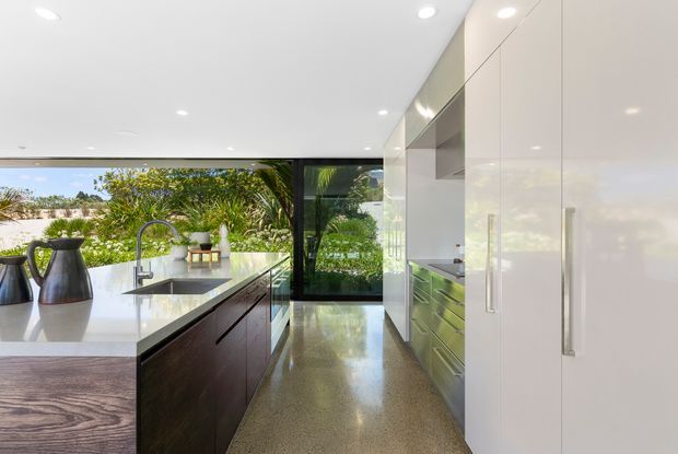A bright, contemporary kitchen with a large central island, dark timber cabinetry, white stone benchtops, integrated white floor-to-ceiling cabinetry, and stainless-steel appliances. Full-height black-framed glazing opens views toward landscaped greenery, while polished concrete flooring and recessed ceiling lights create a clean, refined finish.