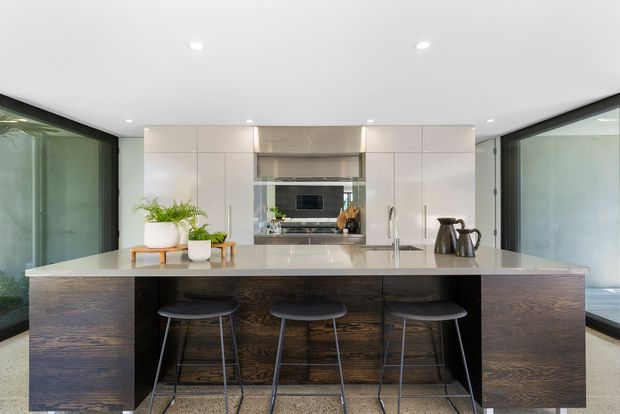Bright contemporary kitchen with a large central island and three black stools. The island has a dark timber-look base and a pale stone benchtop, while the rear wall features sleek white handleless cabinetry, integrated appliances and a stainless-steel rangehood. Full-height dark-framed glazing on both sides brings in abundant natural light and creates a clean, open feel.