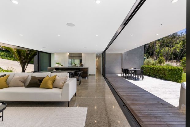 A bright, open-plan contemporary living and dining area with full-width retractable glazing opening onto a timber deck and landscaped garden. The room features polished grey flooring, a white ceiling with recessed downlights, a large white sectional sofa with neutral and mustard cushions, and a compact kitchen with dark cabinetry and a central island. An outdoor dining setting is visible on the deck beyond the open glazing.