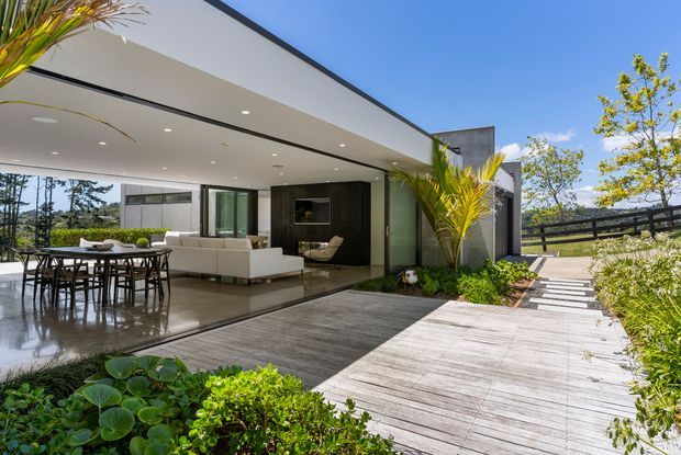 A contemporary single-storey house with a low flat roof and extensive openable glazing, viewed from a landscaped timber deck and garden path. The covered outdoor living area contains a dining table and chairs, while a pale sofa and dark built-in joinery are visible inside. White rendered surfaces, dark window frames, timber decking, concrete-toned paving, palms and clipped planting create a refined indoor-outdoor setting.