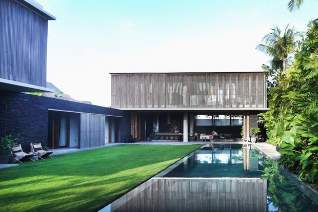 Contemporary tropical residence arranged around a long rectangular swimming pool and manicured lawn. The house features dark stone and vertical timber cladding, large glazed openings, a projecting upper-level box, and lush palm landscaping in bright daylight.