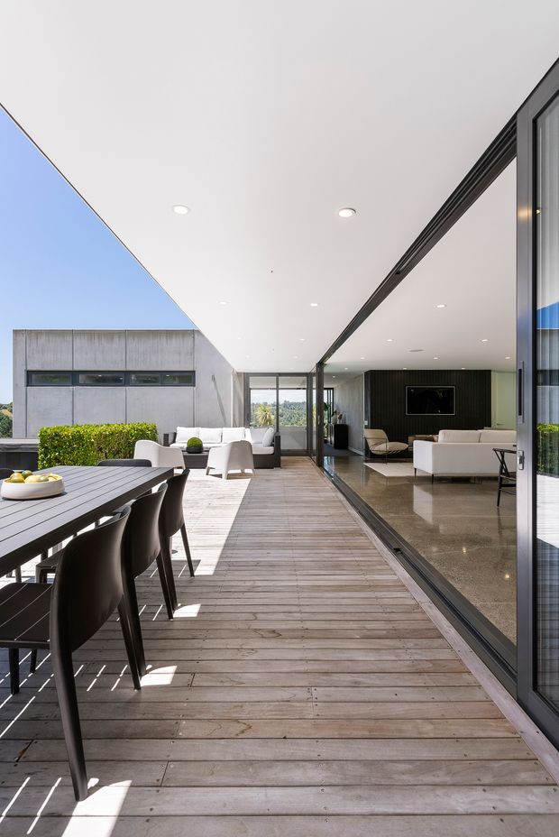 A bright contemporary indoor-outdoor living area opening onto a spacious timber deck. The deck includes a long dark dining table with black chairs, while the glazed interior reveals a refined living room with pale sofas, polished stone flooring, dark vertical wall panelling, and extensive floor-to-ceiling sliding glazing. A large white projecting ceiling creates a sheltered terrace and strong geometric lines.