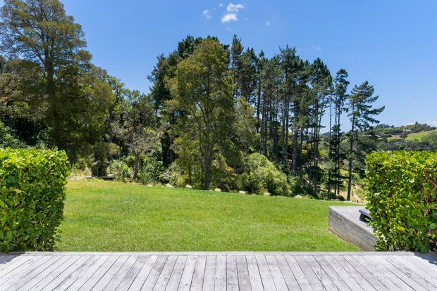 A sunny landscaped outdoor area viewed from a broad timber deck. The scene includes a neatly mown lawn, clipped hedges, mature trees, dense planting, and a raised timber-edged garden feature or seating area at right. The image has a bright, natural suburban setting with distant residential landscape visible through the trees.