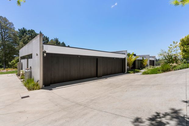 Exterior view of a contemporary low-profile residential property, dominated by a wide double garage with dark vertical-panel doors and a pale rendered or concrete facade. The building sits on a broad sloping concrete driveway, with landscaped planting and mature trees around the site. Flat rooflines, crisp geometric forms, and integrated outdoor lighting create a minimalist modern appearance under bright clear daylight.