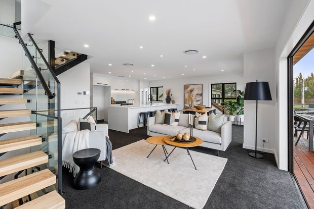 Bright contemporary open-plan living room with a white kitchen in the background, a large pale sectional sofa, round timber coffee tables, light rug, dark carpet, and extensive glazing opening to a timber deck. A floating timber-and-glass staircase with black metal supports occupies the left side, with a black pendant-style floor lamp and indoor plants adding contrast.