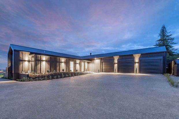 Twilight exterior view of a low, modern L-shaped residence or garage wing with dark metal cladding, extensive floor-to-ceiling glazing, warm interior lighting, and multiple wide garage doors. A broad paved driveway dominates the foreground, with restrained landscaping and a tall evergreen tree at the right.