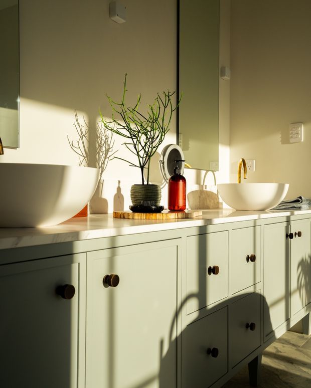 A warmly sunlit bathroom vanity with pale green shaker-style cabinetry, a light stone benchtop, two white vessel basins, and brass wall-mounted taps. Large mirrors reflect the cabinetry and decorative branches, while dark round cabinet knobs, a red soap dispenser, and sculptural accessories add contrast. Strong late-afternoon shadows create a calm, refined atmosphere.