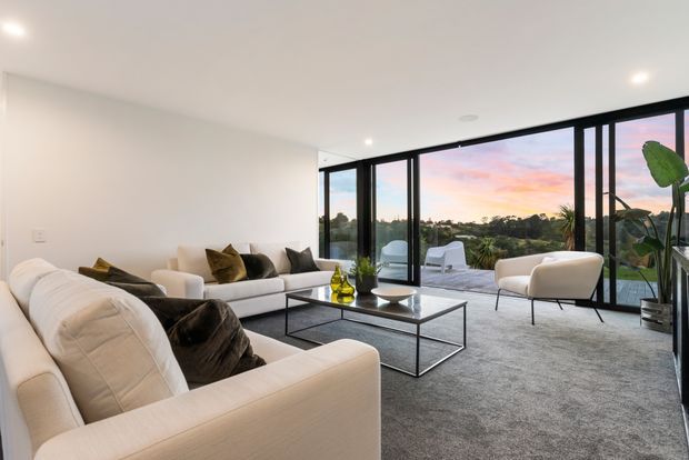 Bright contemporary living room with large cream sectional sofas, a dark metal-framed glass coffee table, and neutral carpet. Full-height black-framed glazing opens the room to a sunset outlook, with a pair of light lounge chairs and indoor plants near the windows.