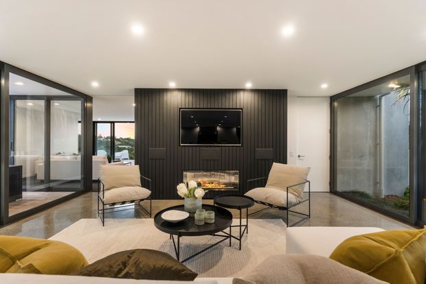Contemporary living room with a black vertical timber-slat media wall, built-in fireplace, and television. The room has polished concrete flooring, neutral furnishings, black-framed glazing and doors, recessed ceiling lights, and a glazed courtyard or planted lightwell visible at right.