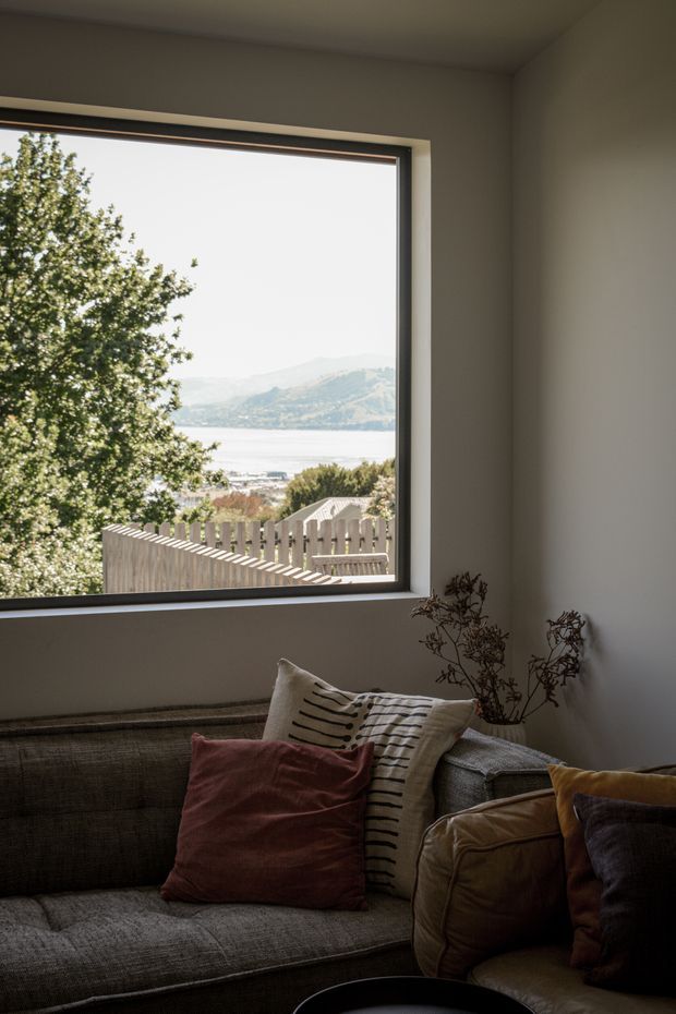 A cozy living room corner with a large picture window overlooking a lake, shoreline, and mountainous landscape. A textured grey sectional sofa fills the foreground, layered with rust, cream, mustard, and dark cushions. A small vase of dried branches sits beside the sofa, and the room has plain light walls with a dark-framed window.