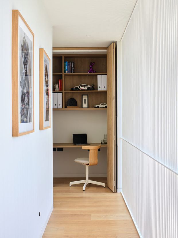 A compact, bright built-in home office nook viewed from a hallway. The room features a timber desk and shelving unit with books, folders and decorative objects, a light upholstered swivel chair, pale timber flooring, white walls, framed artwork, and a white vertically ribbed sliding or folding screen at the right. The space has clean contemporary joinery and abundant soft daylight.