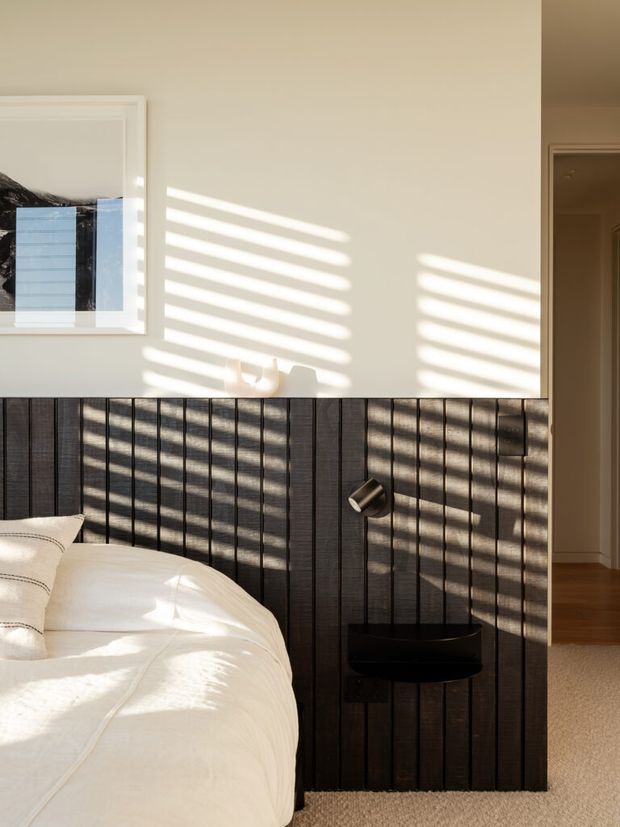 Sunlit bedroom interior featuring a partially visible bed with white bedding, a dark vertically slatted timber feature wall and integrated black bedside shelf with reading light. Warm neutral walls, timber flooring, carpet, framed artwork, and strong striped sunlight from window blinds create a calm, contemporary atmosphere.