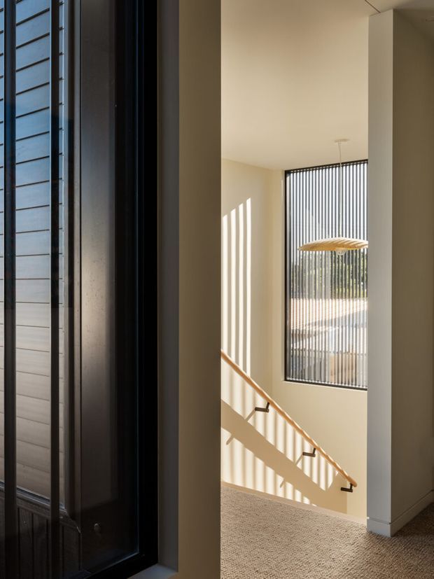 A sunlit interior stairwell or landing viewed from an entry area, with warm off-white walls, a carpeted floor, timber handrail, and a tall window screened by closely spaced vertical dark metal elements. Strong linear shadows fall across the walls and stairs, while a simple woven pendant light hangs in the double-height space. A dark vertical-slatted exterior or entry door is visible in the foreground.