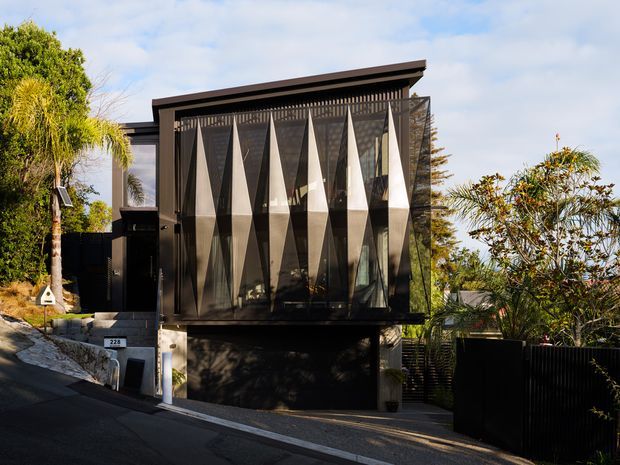 Street-facing exterior view of a contemporary multi-level residence on a sloping suburban site. The dark facade features a projecting upper volume with full-height glazing screened by a striking geometric vertical fin or folded-panel facade. A recessed garage occupies the lower level, with a narrow driveway, boundary fencing, and landscaped planting around the home.