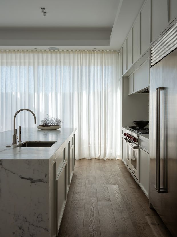 A bright, refined contemporary kitchen viewed along a narrow galley-style layout. A large white marble island with integrated sink and brushed metal gooseneck faucet occupies the foreground, while pale grey built-in cabinetry, a stainless-steel refrigerator, cooktop, and wall oven line the right side. Full-height sheer white curtains diffuse daylight at the far end, and light timber flooring runs throughout.