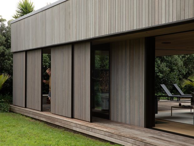 Exterior view of a contemporary single-storey pavilion-like building clad in vertically ribbed timber boards, with large dark-framed glazed openings and sliding timber screens. A timber deck extends through the open living area toward a landscaped garden with outdoor lounge furniture and palms.