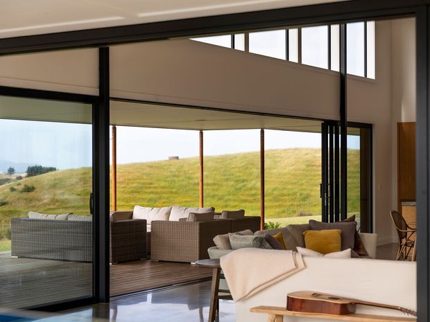 A contemporary living room with expansive floor-to-ceiling glazing opening onto a covered outdoor deck. The interior includes a large light-coloured sofa, armchair, low timber table, cushions, and a guitar, while wicker outdoor seating is visible beyond the open glazing. A grassy rural landscape is visible outside.