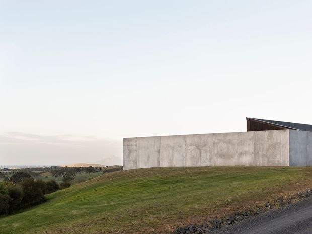 A minimalist contemporary building set into a gently sloping grassy site. The structure features a long, raw-looking concrete wall and a dark angled roof form, with expansive rural farmland visible beyond. The composition emphasizes the building’s low horizontal massing and integration with the landscape.