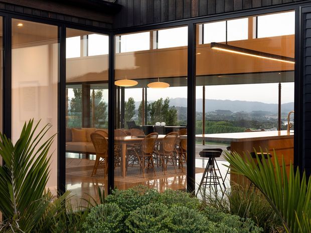 Contemporary indoor-outdoor living and dining space viewed through large black-framed glazed doors. The room features a timber dining table with woven chairs, a lounge area, polished concrete or stone flooring, and a compact kitchen or bar with a timber-clad island. Warm pendant and linear lighting contrast with dark vertical timber wall cladding. Dense planting in the foreground and a distant mountainous landscape reinforce the connection to the outdoors.