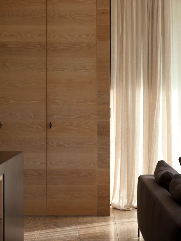 A warm, minimalist interior detail showing full-height timber veneer cabinetry or wall panelling with concealed doors and a small dark handle. Floor-to-ceiling sheer curtains admit soft daylight beside a dark upholstered sofa, while large polished beige stone or porcelain floor tiles reflect the light. The composition emphasizes clean vertical lines, natural wood grain, and a calm neutral palette.
