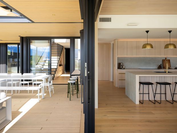 Bright open-plan residential interior showing a dining area and contemporary kitchen connected by a large black-framed glazed opening. The space features extensive pale timber ceiling and flooring, a long pale timber kitchen with integrated cabinetry, a light stone island, ribbed island detailing, black bar stools, pendant lights, and a staircase beyond the dining area. Strong daylight enters through large windows and sliding doors, creating a spacious indoor-outdoor connection.