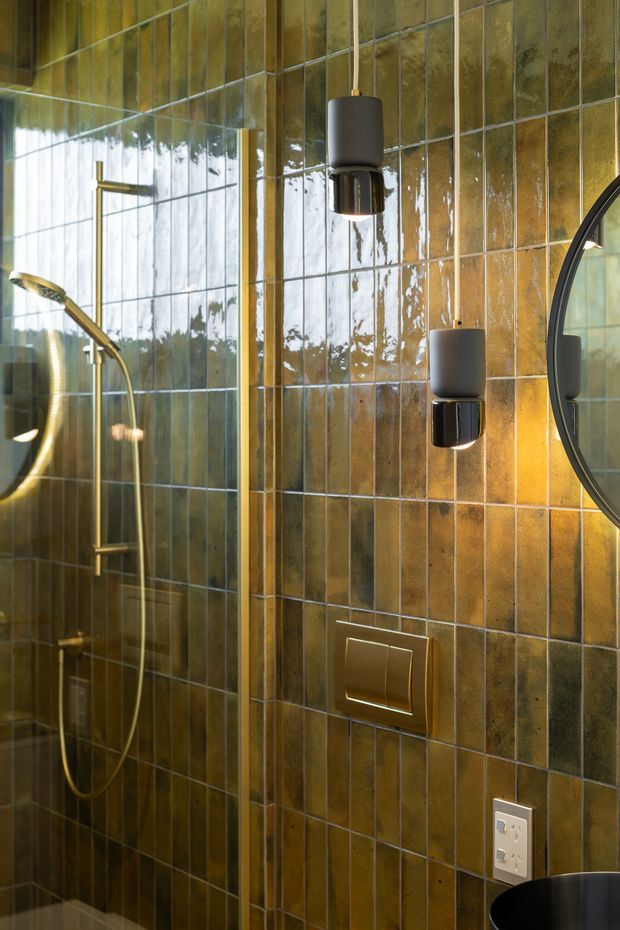 Close-up view of a richly finished bathroom shower area with glossy, vertically arranged olive-brown rectangular wall tiles, brass shower hardware, a glass shower screen, cylindrical wall lights, a flush switch, and a partial round mirror and basin at the edge of the frame. The reflective tile surface creates a warm, moody atmosphere.