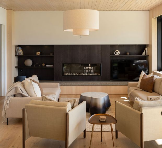 Warm, refined living room with a custom full-width media and fireplace wall. The dark timber feature joinery incorporates open shelving, a linear fireplace and television, balanced by pale upholstered sofas and armchairs. Light timber flooring and a timber-lined ceiling create a cohesive natural palette, while a large sculptural pendant provides a focal point.