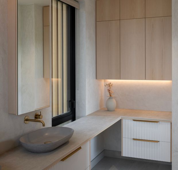 A refined contemporary bathroom vanity area with a floating stone-look benchtop, vessel basin, wall-mounted brushed brass tapware, large mirrored cabinet, and integrated light-wood cabinetry. Vertical fluted drawer fronts and warm under-cabinet lighting add texture, while a narrow black-framed window brings daylight into the compact space.